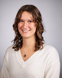 A woman with long brown hair wearing a white sweater in front of a grey backdrop.