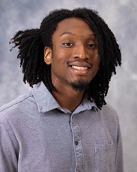 A man with dark hair and a grey shirt smiles in front of a grey background.