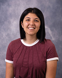 A woman in a maroon shirt and dark hair in front of a grey background.