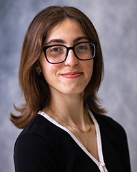 A woman in glasses, dark hair, and a dark shirt in front of a grey background.