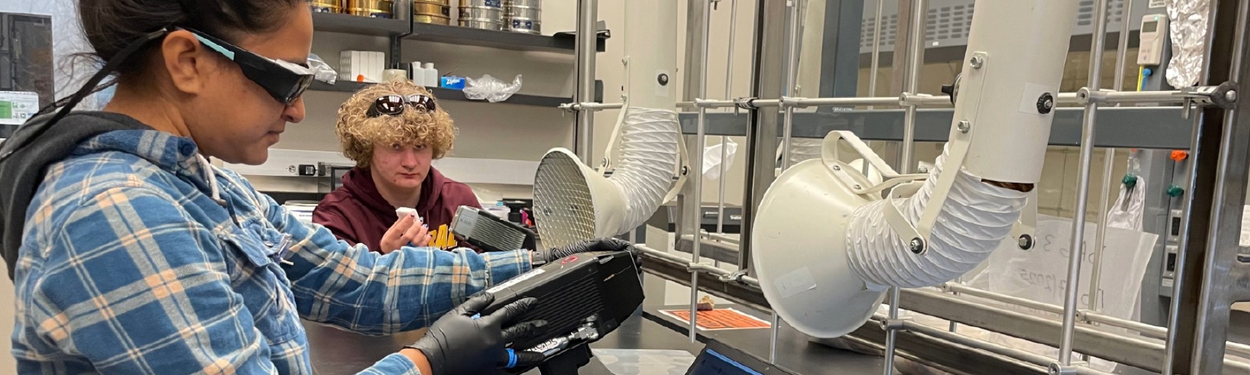 two biology students in a lab setting wearing black goggles and operating equipment for a project