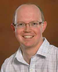 John Allen wears a button up shirt and smiles for a headshot.