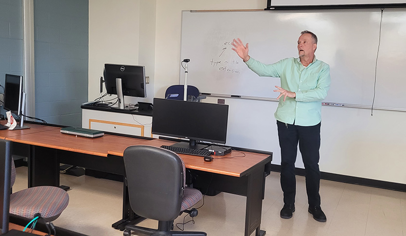 Geography faculty member in dark pants and a green shirt in front of a whiteboard in a classroom.