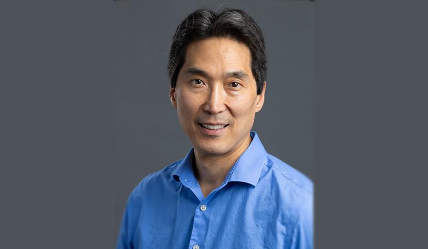 A man with dark hair and a blue shirt in front of a grey background.