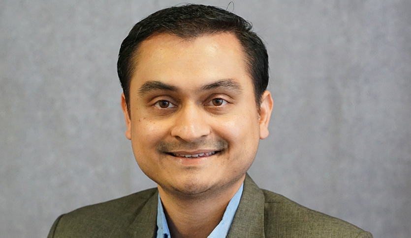 Nirajan Budhathoki in a blue shirt and brown jacket against a grey background smiling at the camera.
