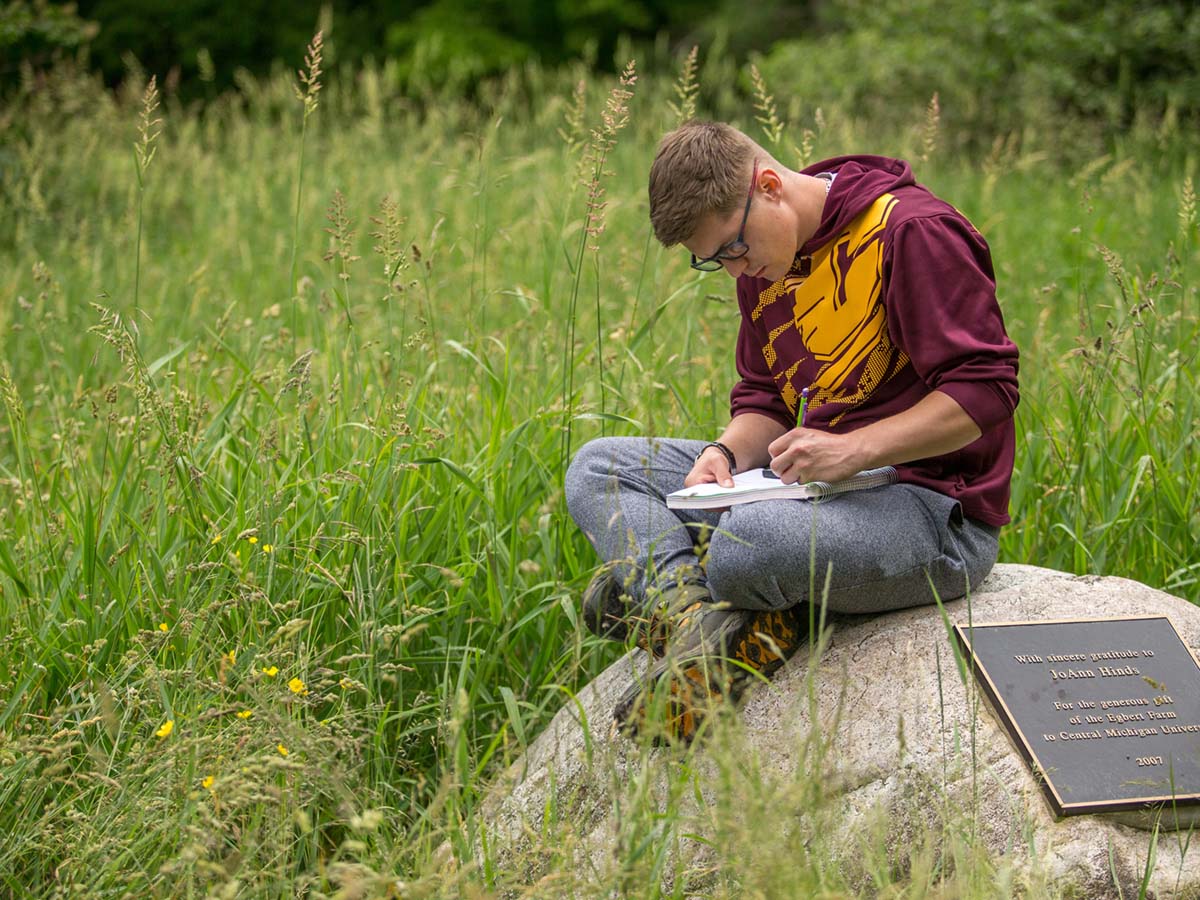 Student sitting on a rock in a field writing notes.