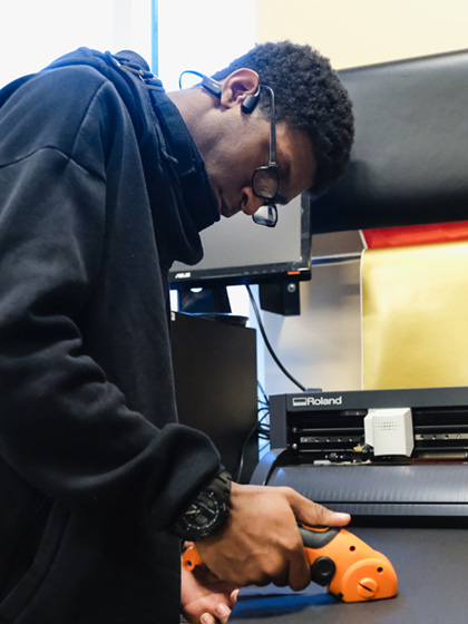 InSciTE student in a dark sweatshirt and glasses cutting material in an InSciTE classroom.