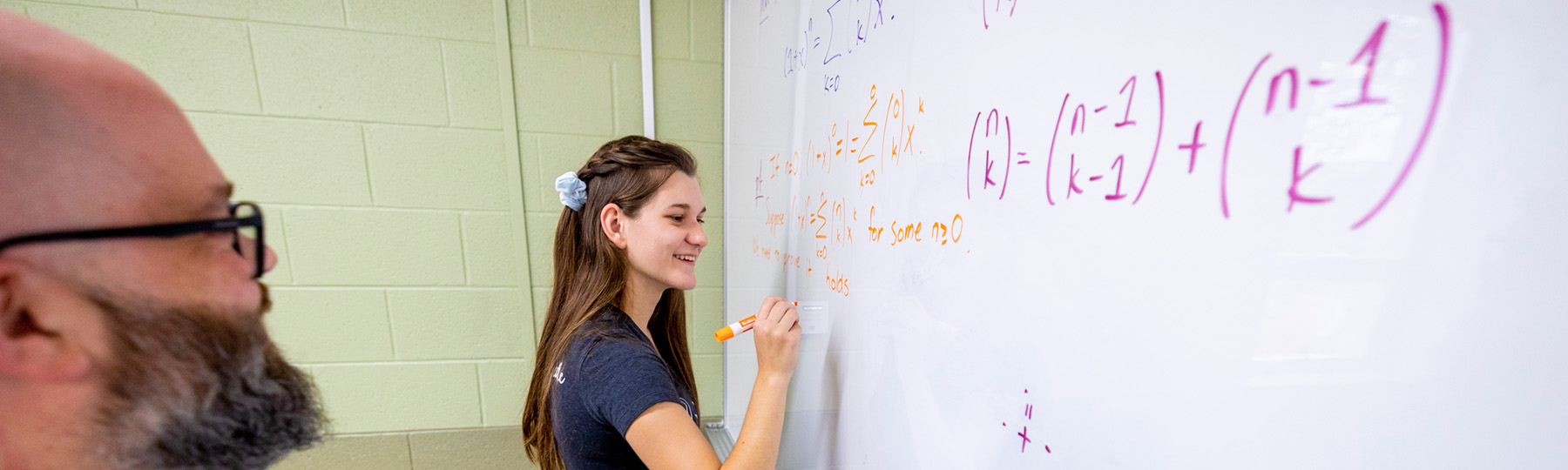 A student in a blue t-shirt completing a mathematics equation on a white board while being observed by a mathematics faculty member.