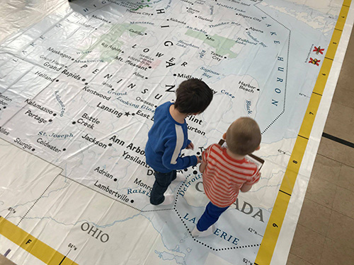 Two young elementary students standing, one in a blue shirt and one in a red a white striped shirt,  on a giant map of michigan.