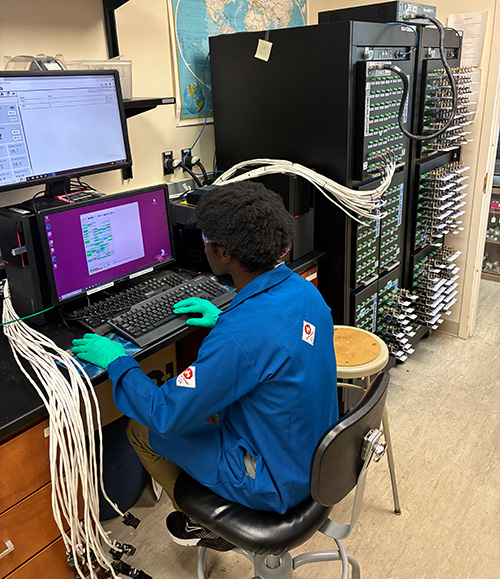 20250701_IsaacBoakya A man in a blue lab coat sitting at a computer that is connected with a large bundle of wires to a large rack of equipment.