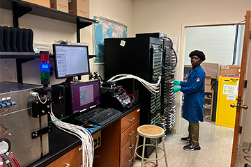 A student in a blue lab coat standing in front of 2 tall racks of equipment that are connected to a computer.