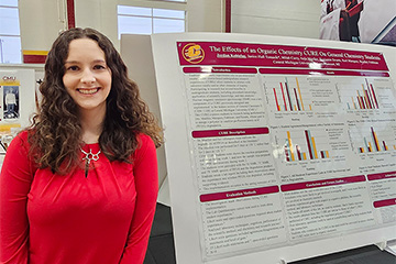A student in a red shirt and long dark hair standing next to her research poster.