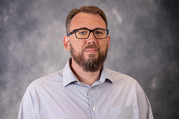 A man with glasses, a beard, and button up shirt in front of a blue background.