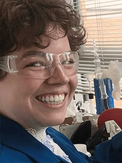 a woman wearing safety glasses in a lab, smiling at the camera