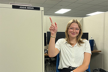 A woman in a white shirt and glasses pointing up toward a sign that says her name, Jessie Servis.