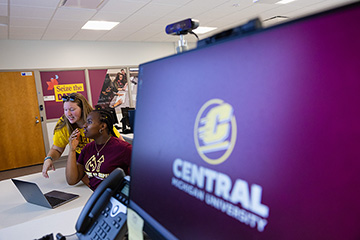Two students looking at a laptop screen with a computer screen in the foreground that says Central Michigan University on it.