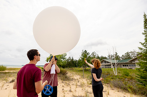 Three students on a sandy beach holding a large white weather balloon