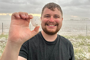 Student stands near a body of water holds up a small white spherical sample and smiles