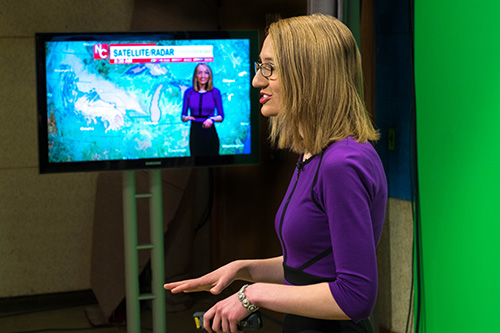 A woman in a purple sweater with glasses standing in front of a green screen.