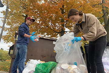 Eric Urbaniak and another student sorting through trash bags next to a dumpster on a fall day on campus.