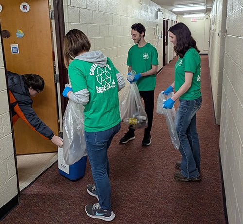 Three students in green t-shirts holding bags of recycling are accepting items from a student in their residence hall room.