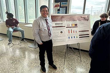 A student in a shirt and tie standing next to a research poster.