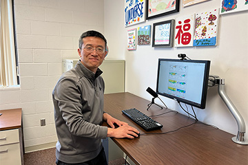 Tao Zheng in a grey shirt standing at a computer desk while smiling at the camera.