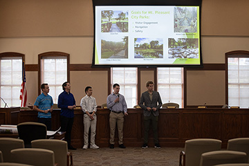 Five students standing in front of a large display screen pitching their ideas for signage withing Mt. Pleasant Parks.