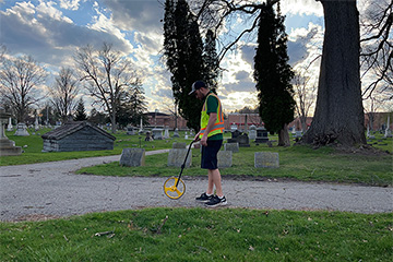 Anthony Kowalchick in a cemetery, wearing shorts and a high visibility vest using a rolling device to measure ground distance.