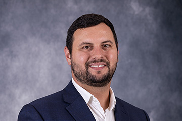A man in a white button up shirt and blue jacket smiles in front of a grey background.