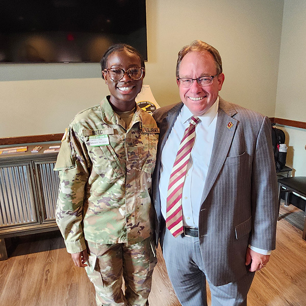 20250910_AutumnForNews A woman in army fatigues and a man in a suit standing together in an office.