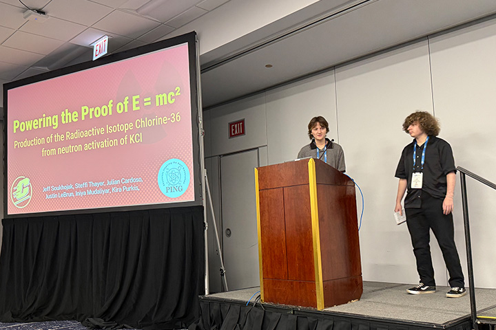 20251117_PINGStory_720x480 Two students stand at a podium during a presentation. a slide is projected on a screen, showing the text Powering the Proof of E equals M C squared