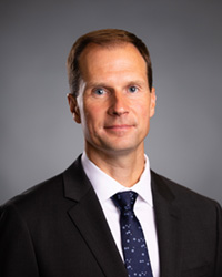 A man in a suit coat, collared shirt and tie poses for a headshot.