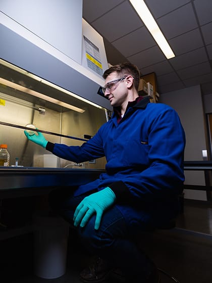 A male student in a blue lab coat and safety goggles holds a petri dish under a hood in a research lab on campus.