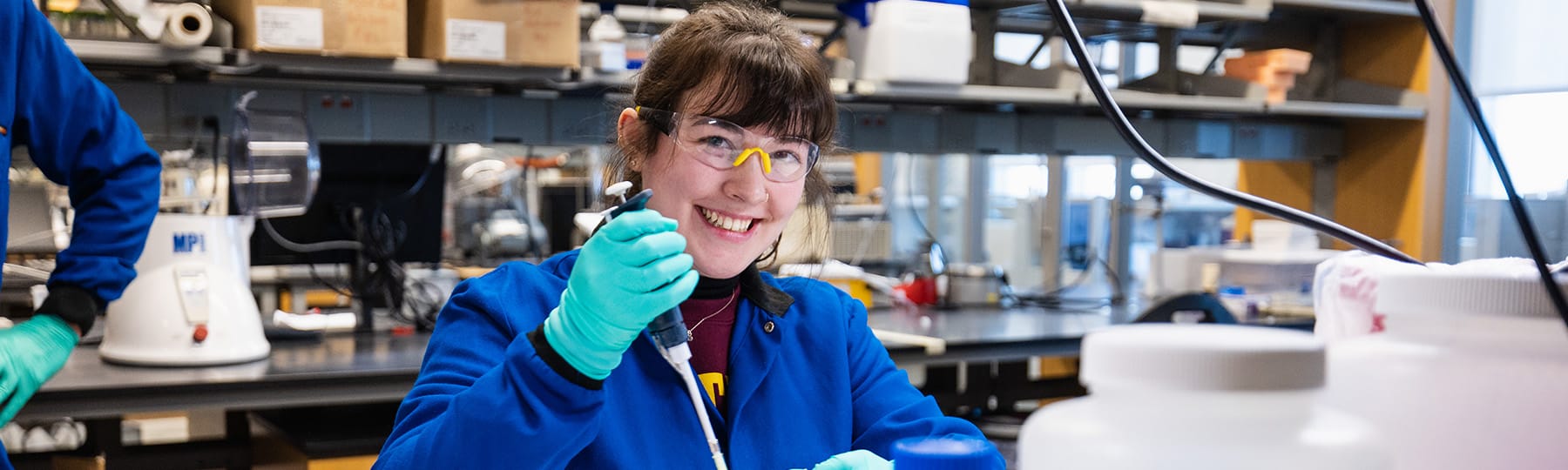 A female student in a blue lab coat and safety glasses holding a pipette works in a lab on campus.