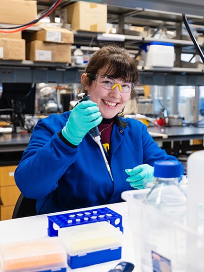 A female student in a blue lab coat and safety glasses holding a pipette works in a lab on campus.