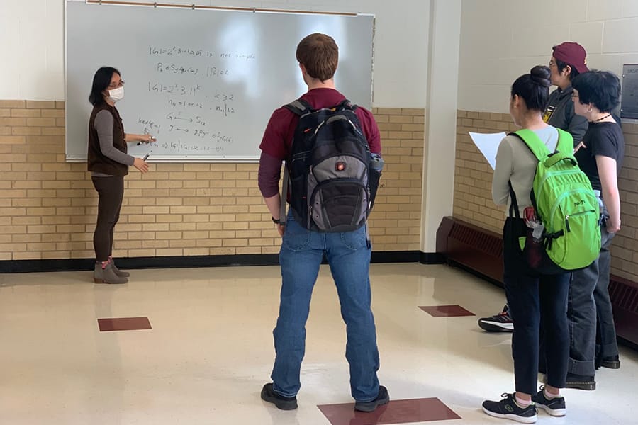 An an instructor and group of mathematics students stand in front of a whiteboard with an equation written on it.