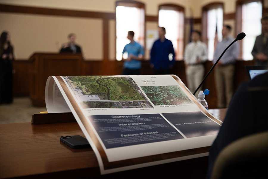 A close up of a map of Mount Pleasant parks sitting on a desk. In the background a student presents at a podium.