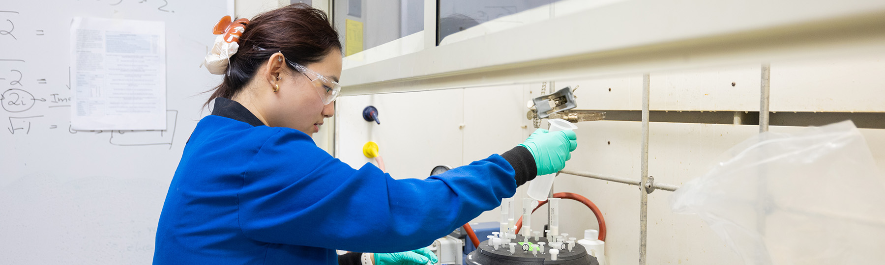 A woman wearing goggles, a blue lab coat, and gloves holding a test tube.