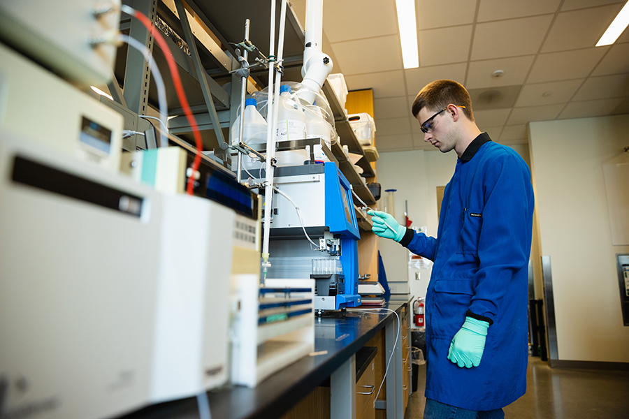 Male student stands in front of lab equipment wearing protective gloves, coat and glasses.