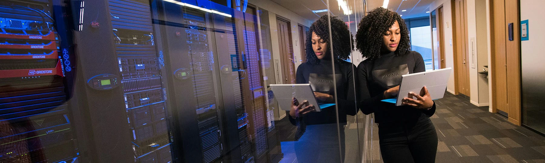 A woman with knowledge in computer science holds a laptop and leans against a glass panel on the outside of a server room