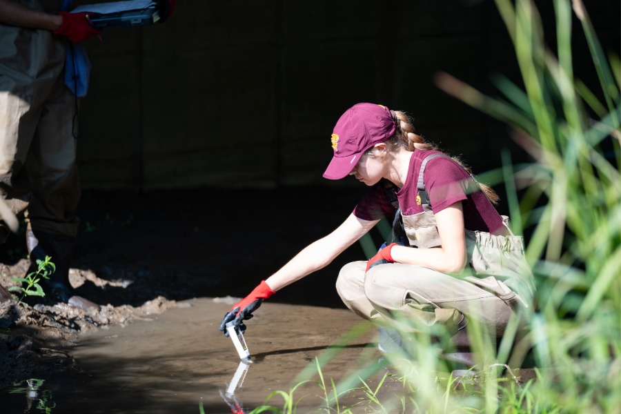 a CMU student wears overalls and high rubber boots as she kneels in a creek to collect a water sample with lab equipment