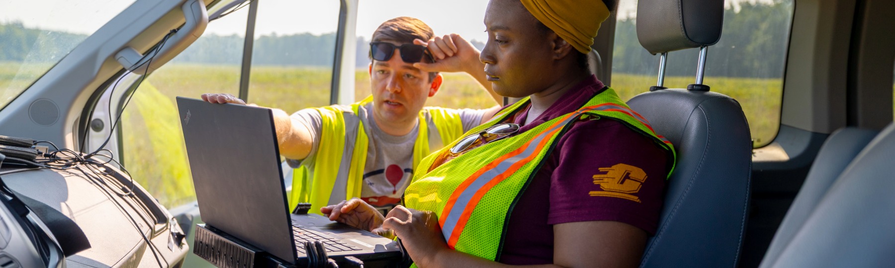 a CMU student and faculty discuss findings while looking at a laptop inside a car in a grassy field