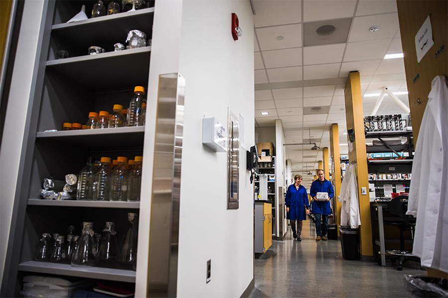 A student and faculty member wearing blue lab coats walk down the hallway of a science lab.
