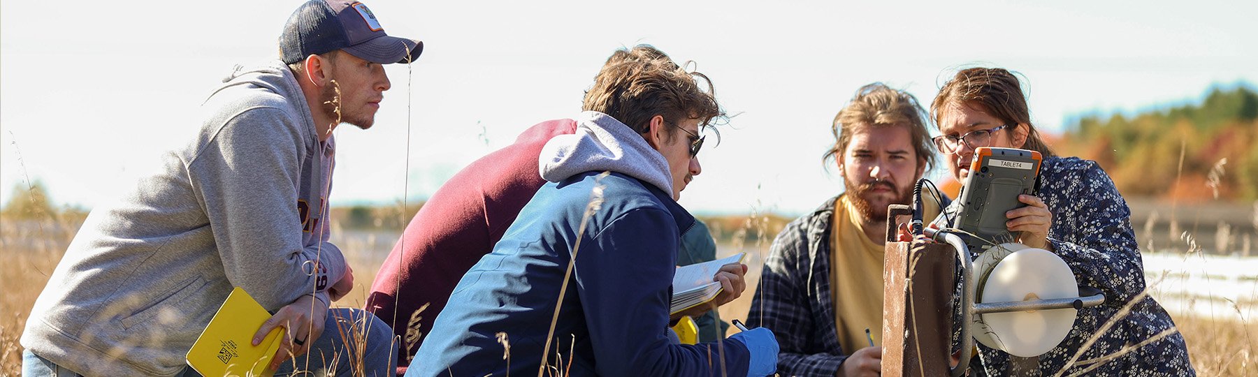 A professor and group of students gather around a ground water measurement instrument in a field.