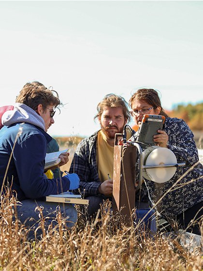 A professor and group of students gather around a ground water measurement instrument in a field.