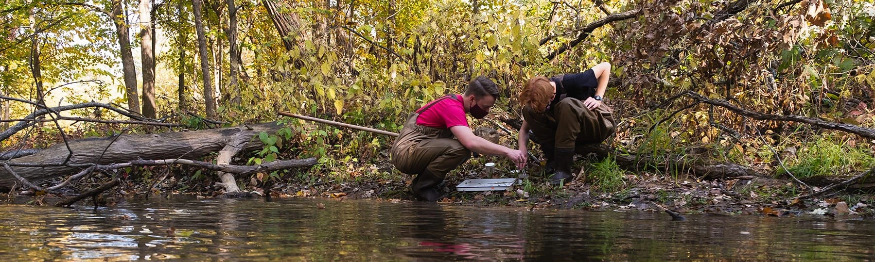 Two student wearing waders crouch on the bank of a river examining it.