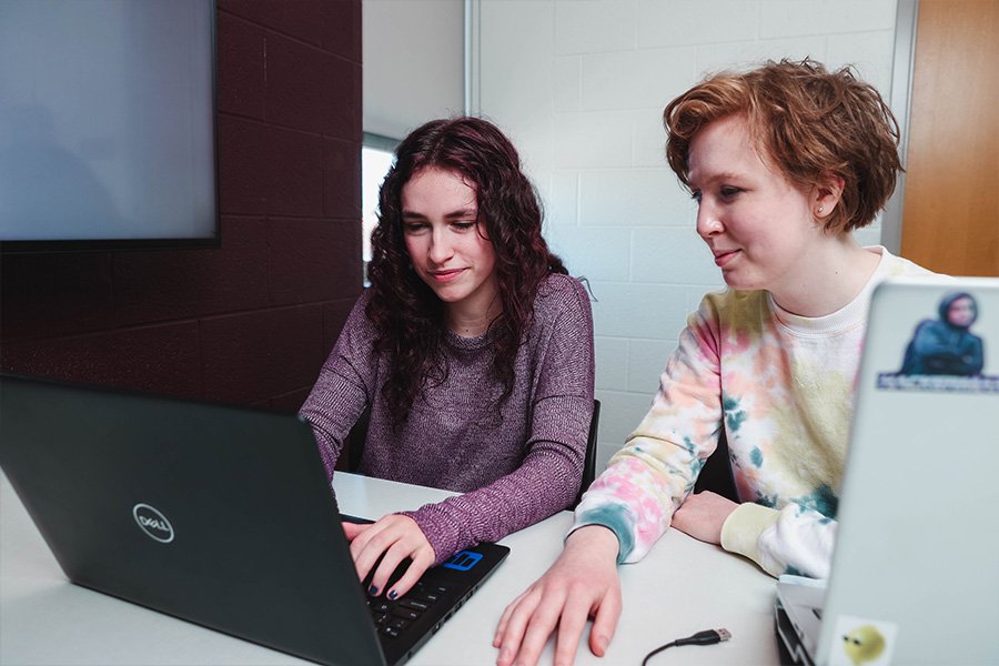 Two computer science majors sit at a desk, collaborating behind laptop computers.