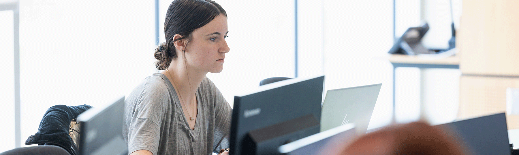 A student wearing a gray shirt, sitting in a classroom at a desk, looking forward at a computer screen.