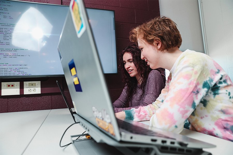Two students work on computers next to each other with coding displayed on a screen in the background.